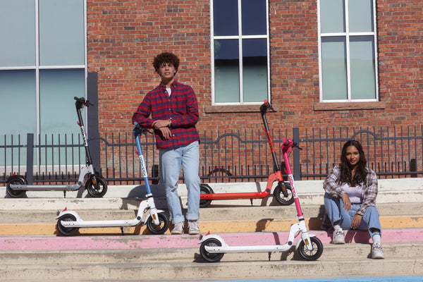 A man and woman with, from Left to Right, a GOTRAX Silver, Blue, Black/Red, and Pink Apex LE Foldable Electric Scooters for Adults