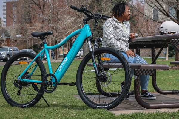 A woman sitting on a park bench next to a GOTRAX Ocean Teal Emerge Electric Bike for Adults with Front Wheel Suspension