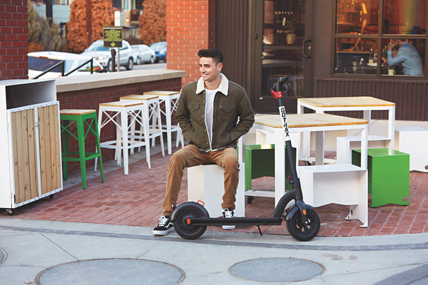 Man sitting at a an outdoor table with the GOTRAX Black G3 Commuter Electric Scooter for Adults with the kickstand down