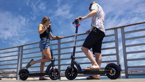 Man on a GOTRAX Black G4 Electric Scooter for Commuters and a Woman on a GOTRAX Black GMAX Ultra Electric scooter for Commuters looking from a pier